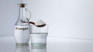 A glass bottle and glass of vinegar with a spoon of baking soda representing the query: How to get transmission fluid out of clothes with vinegar?