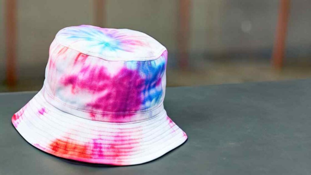 A white colored tie dyed bucket hat with differnt color shades representing the queries: How to tie dye a bucket hat ? Or Is bucket hat tie dye possible ? Or How to tie die bucket hat? Or How to tie dye bucket hats ? Or How to tye dye bucket hats ? What are tie dye hat patterns ?