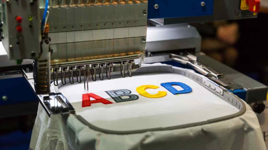 An Embroidery machine sewing a logo of ABCD on a white colored t shirt representing the queries: How to remove logo from shirt ? Or how to remove a logo from a shirt ? Or how to get rid of logos on clothes ? Or How to remove logos from clothing ? Or How to remove logo on t shirt ? Or How to remove nike logo from shirt ? How to remove shirt print ? Or How to remove print fom t shirt ? Or How to remove shirt design ?