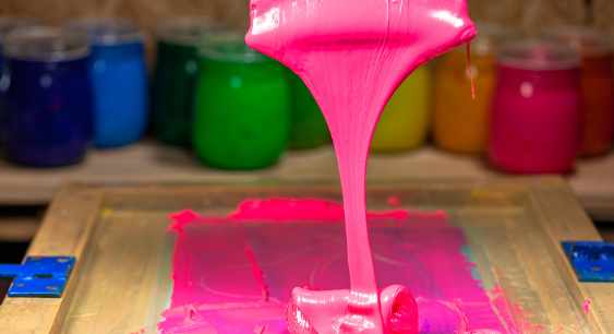 Pink Colored Paint/Ink dripping from the screen printing squeegee with multiple colored inks in the background representing the queries: What is Screen Printing? What are Types of Screen Printing? What is the Process of Screen printing ?