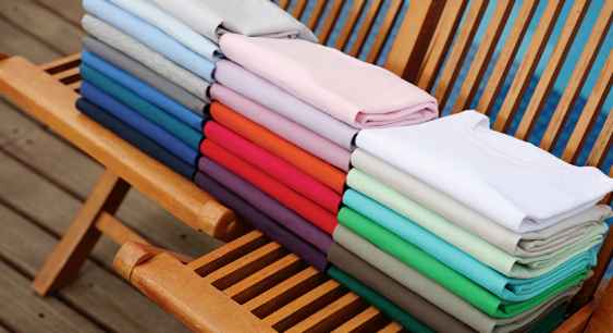 Different colored fabrics of different types pressed and folded on a wooden chair representing the queries: What is a triblend shirt ? Or What is a TriBlend T shirt ? Or What are tri blend t shirts? Or What is a Tri blend t shirt ? Or What is tri blend shirt?
